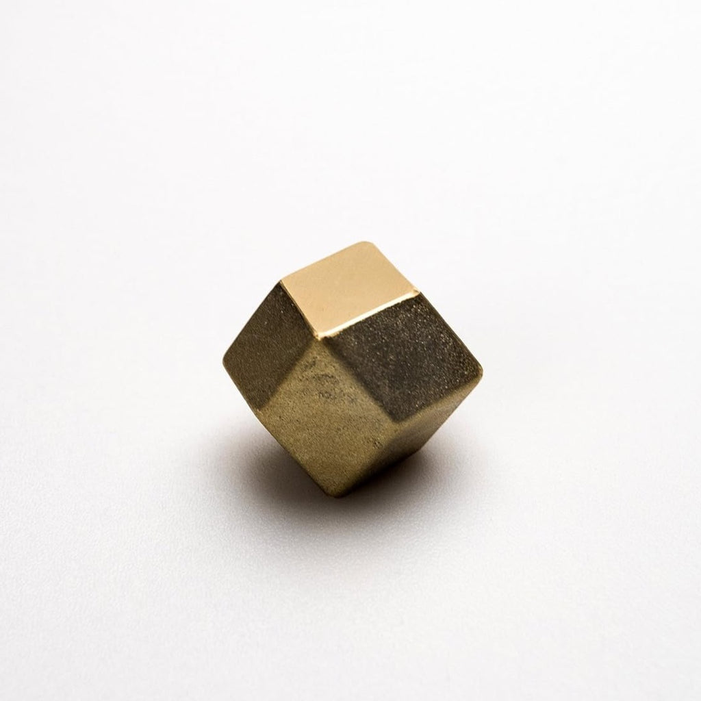 The RHOMBUS PAPERWEIGHT by FUTAGAMI is a metallic golden pyrite cube with a sand-cast brass finish, displaying both rough and smooth textures on a light surface.