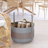 The ELDVARM EMMA WOOD BASKET, featuring a round gray and brown handle, holds firewood and sits on a light wood floor, offering practical storage with stylish design beside wicker and wooden furniture.