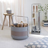 A gray and brown log holder filled with firewood sits beside the ELDVARM EMMA FIRE TOOL COMPANION SET on a carpeted floor, with a candle on a stool and a pillow nearby.
