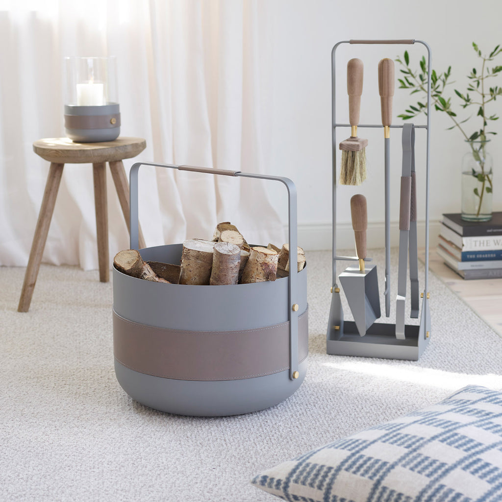 A gray and brown log holder filled with firewood sits beside the ELDVARM EMMA FIRE TOOL COMPANION SET on a carpeted floor, with a candle on a stool and a pillow nearby.