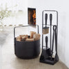 A black EMMA WOOD BASKET holder next to a fireplace made by ELDVARM.