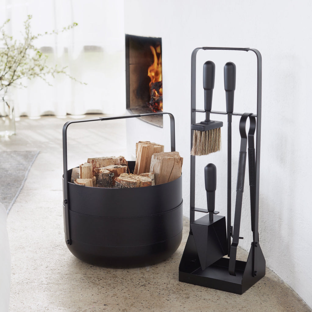 A black EMMA WOOD BASKET holder next to a fireplace made by ELDVARM.