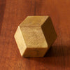 The RHOMBUS PAPERWEIGHT by FUTAGAMI is a small, metallic golden polyhedron with flat faces and a sand-cast brass finish, resting on a wooden surface.