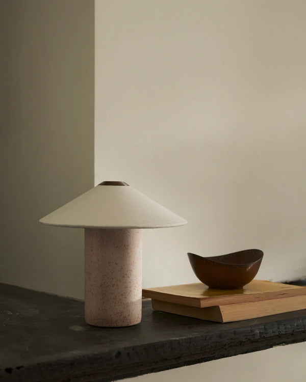 The FUTÉ Table Lamp by LYFA, featuring a Danish design with a wide, flat shade, sits on a dark stone surface next to a small brown oval dish on a wooden block against a plain beige wall.