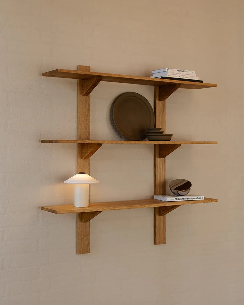 The Danish-designed wooden wall shelves display books, decorative plates, and the sleek FUTÉ Table Lamp by LYFA for an elegant look.