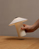 A hand holds the FUTÉ Table Lamp by LYFA, featuring a modern cone-shaped design with a cylindrical base, elegantly illuminating over a wooden surface.