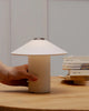 A hand adjusts the LYFA FUTÉ Table Lamp with a conical shade on a wooden table, highlighting its classic Danish design, next to a stack of books.