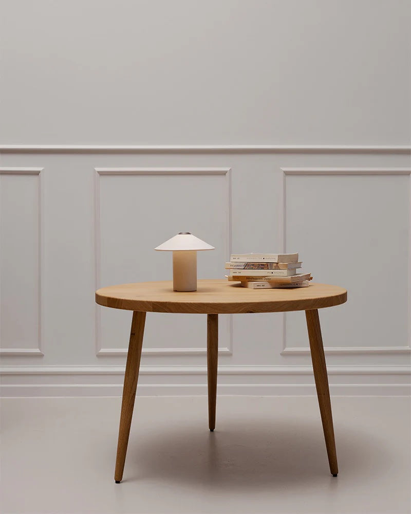 A wooden coffee table elegantly complements a light gray paneled wall and Danish design simplicity, featuring the stylish LYFA FUTÉ Table Lamp alongside stacked books.