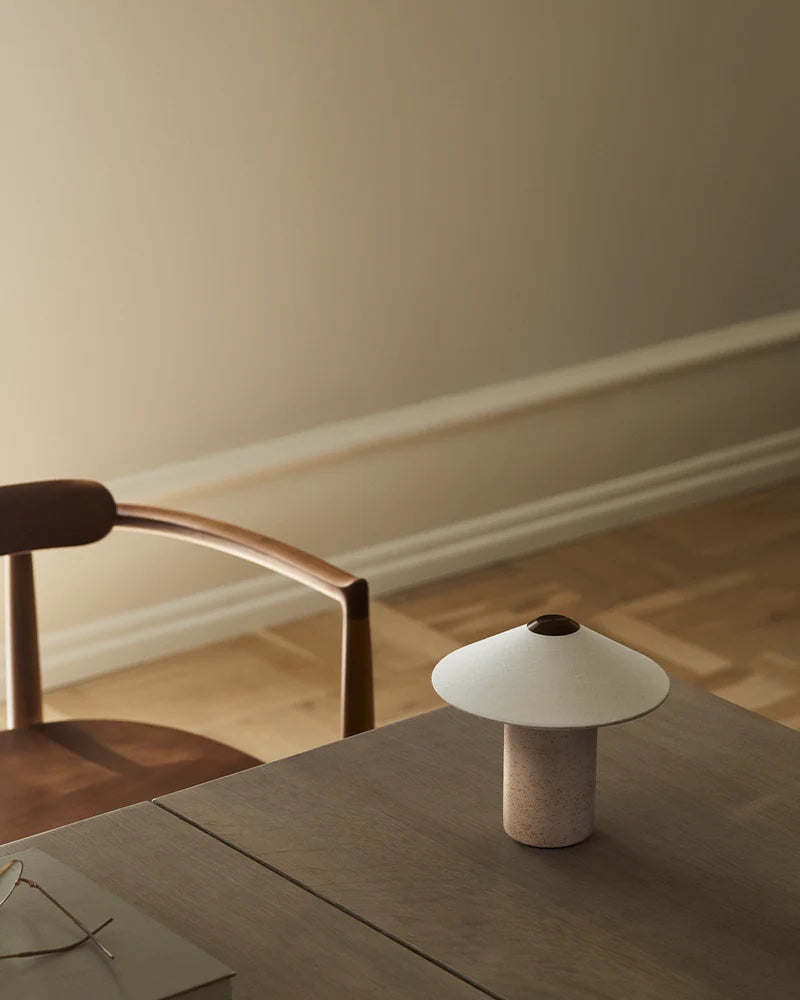 A minimalist interior with a wooden chair and table showcases Danish design. The LYFA FUTÉ Table Lamp, small and white, sits on the table's corner. Neutral-toned walls and floor complete this elegant lighting arrangement.