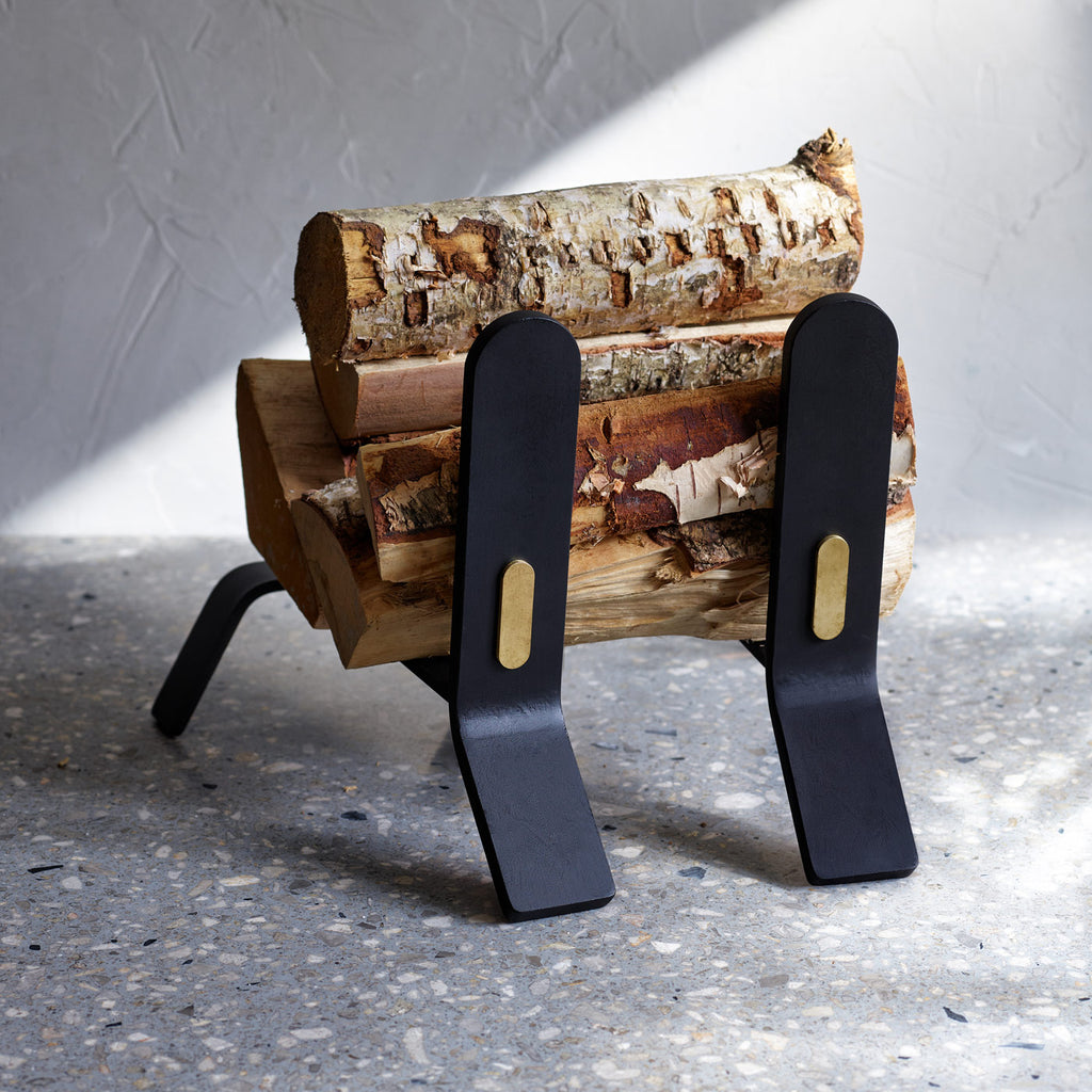 A minimalist black log holder with brass detail, COOPER FIRE DOGS by ELDVARM, on a concrete floor.